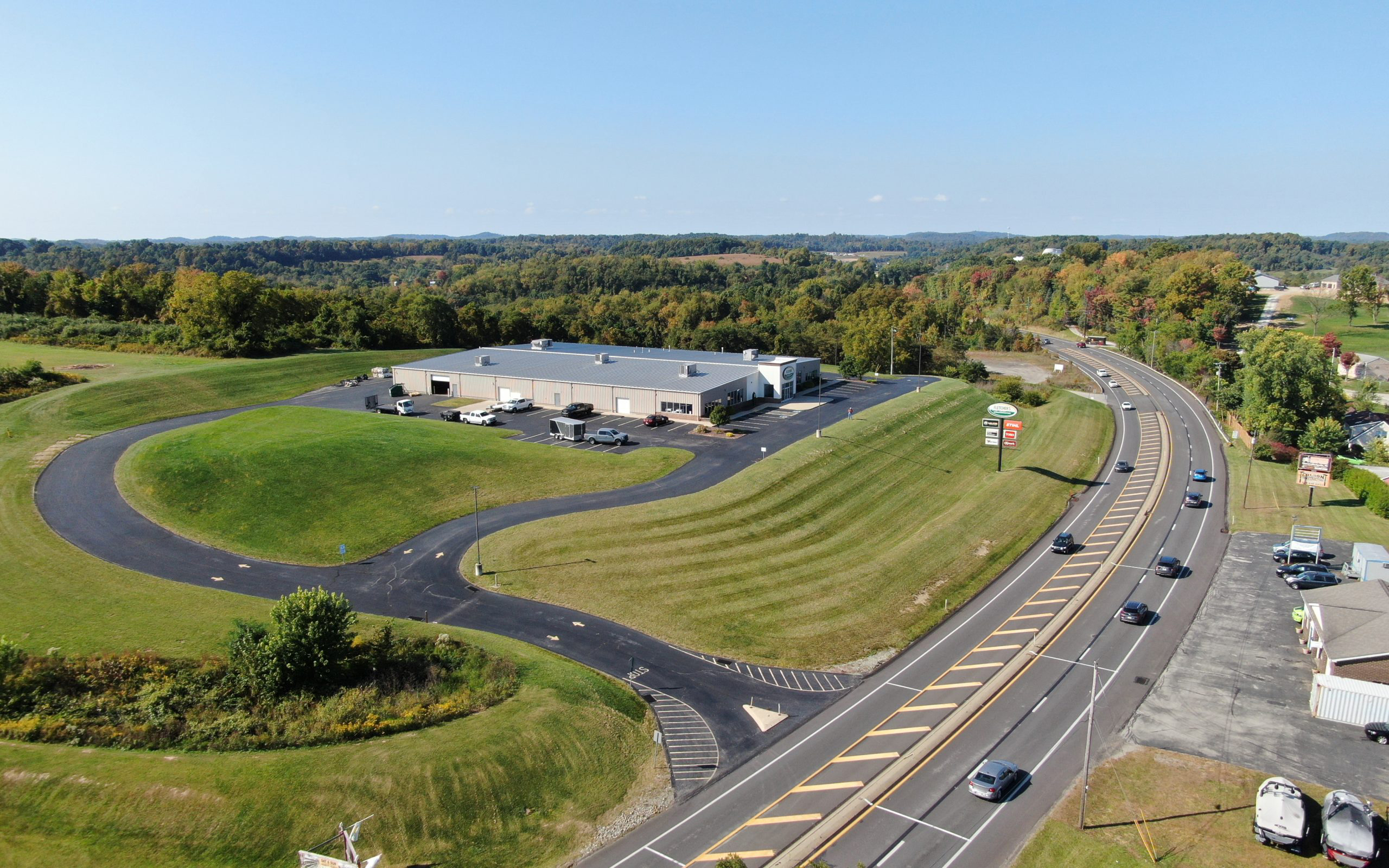 Aerial view of a commercial building beside a winding road, surrounded by green hills and trees under a clear blue sky.